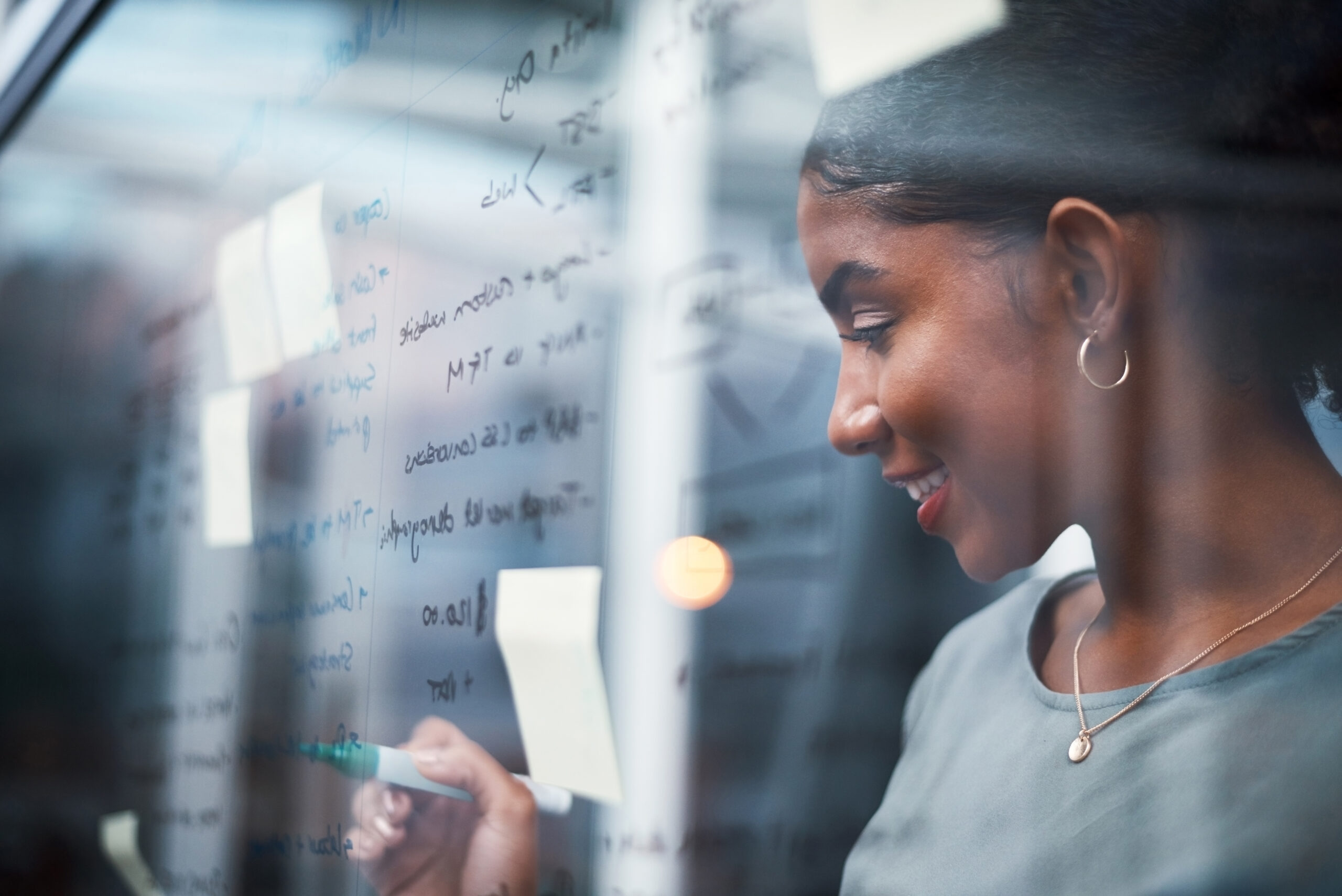 Writing, planning strategy and brainstorming ideas on a glass wall in a modern design office. Young business woman thinking and drawing up a plan for success on a transparent board in the boardroom
